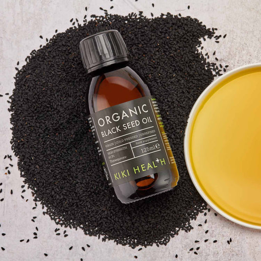 A bottle of organic black seed oil on scattered black seeds next to a bowl of yellow liquid.